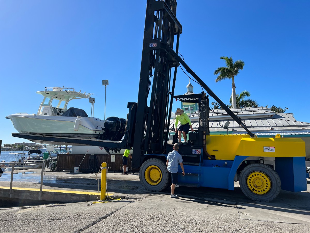 Forklift hoisting a white boat at a harbor with blue sky in the background.