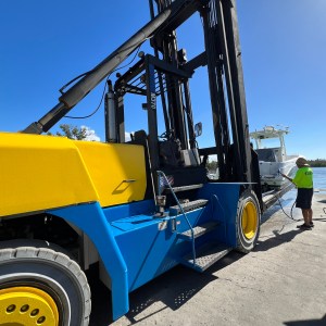 Yellow-blue forklift at dockside, operator fueling a boat under clear blue sky.