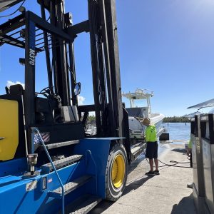 Person in straw hat and neon shirt washes boat next to large blue forklift on sunny day.
